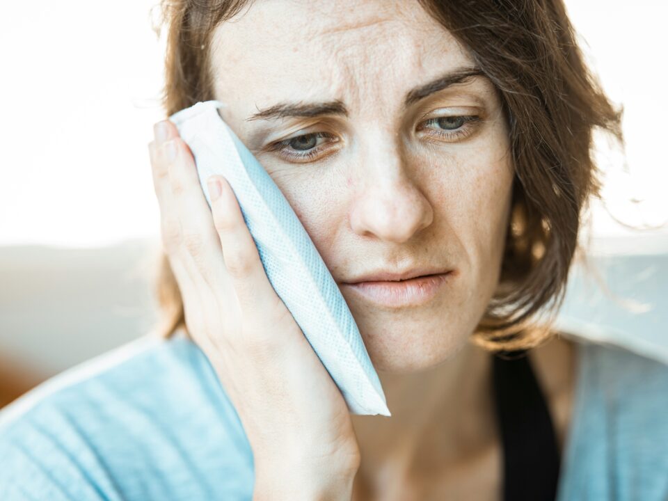 Woman applying cold compress to her jaw, experiencing discomfort, related to TMJ pain relief and chiropractic care at Elevated Chiropractic.