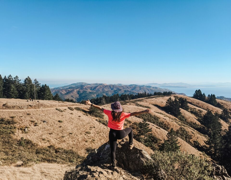 Person standing on a rock with arms outstretched, overlooking a scenic landscape of rolling hills and distant ocean, embodying outdoor wellness and vitality.